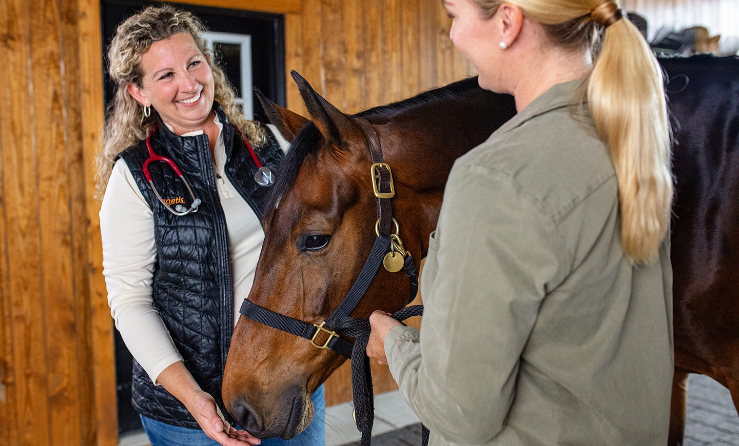 Two women with a brown horse indoors, one smiling with a stethoscope.
